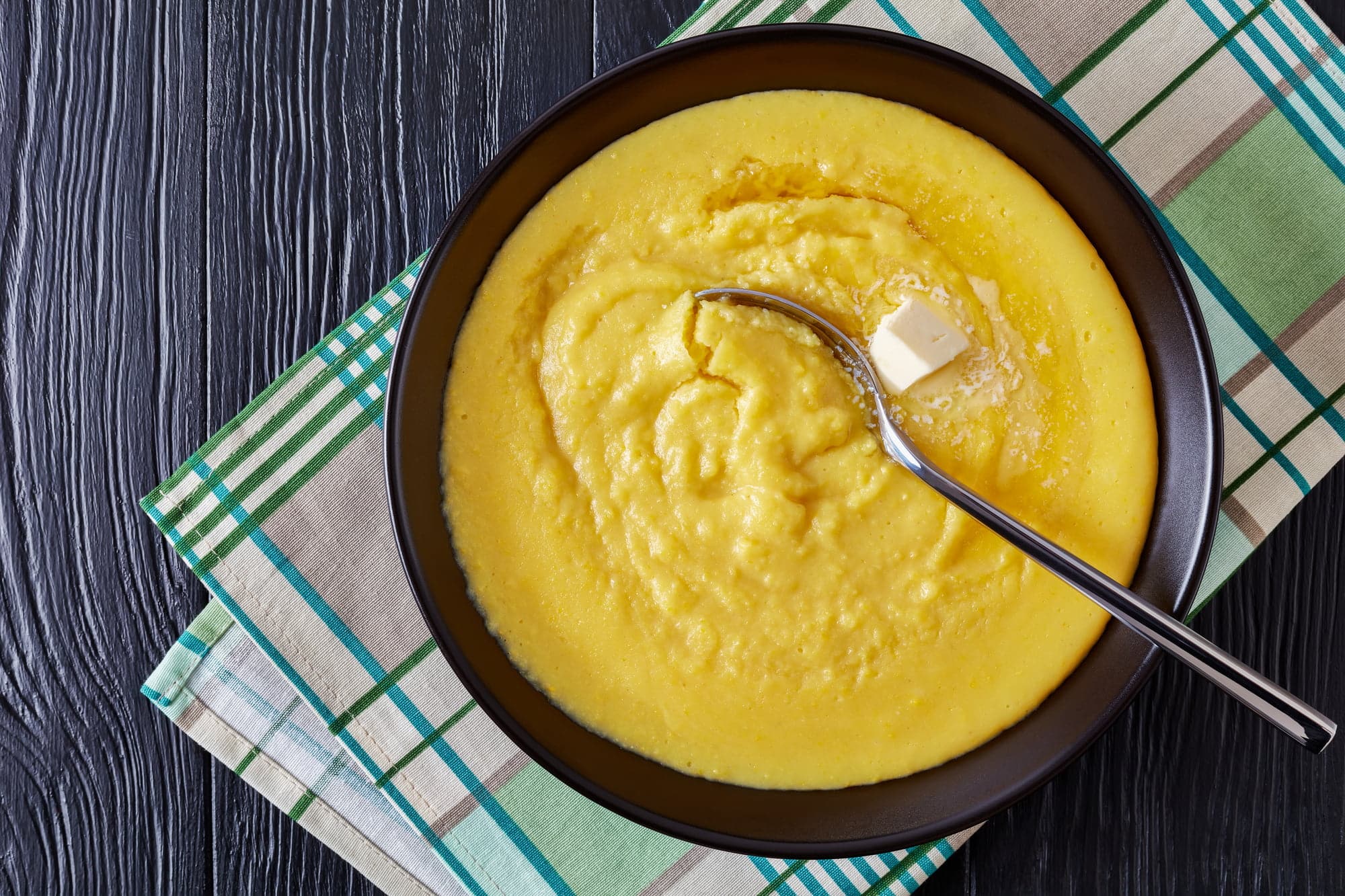 hot cornmeal polenta with melting butter in a bowl with napkin and silver spoon on a black wooden table, horizontal view from above