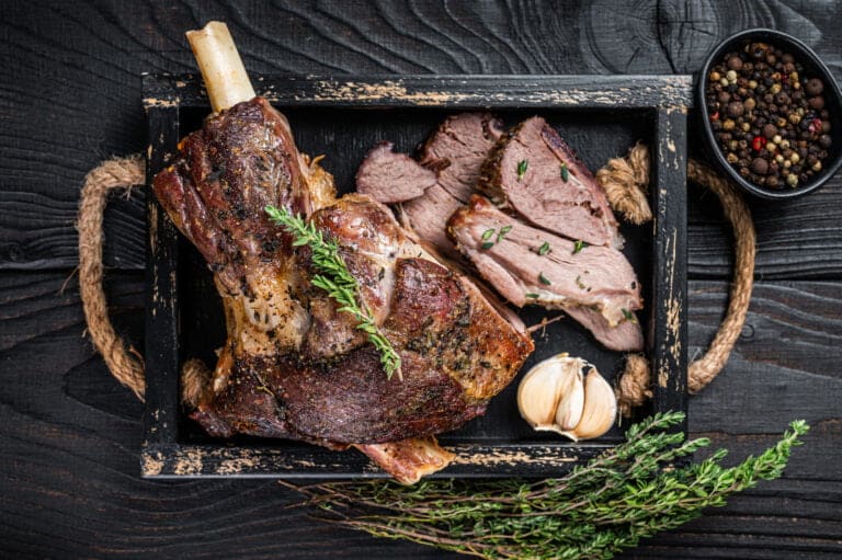 Roasted mutton lamb leg sliced in a wooden tray with meat cleaver. Black wooden background. Top view